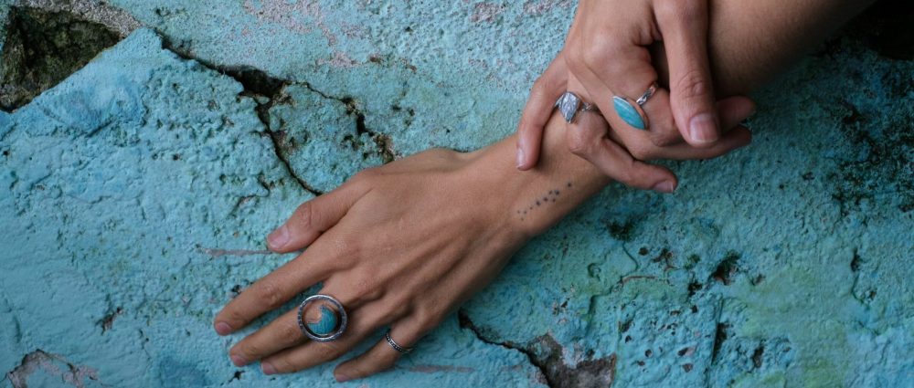 A pair of hands resting on a blue wall. The hands wear wholesale sterling silver rings with moonstone and amazonite genuine gemstones.